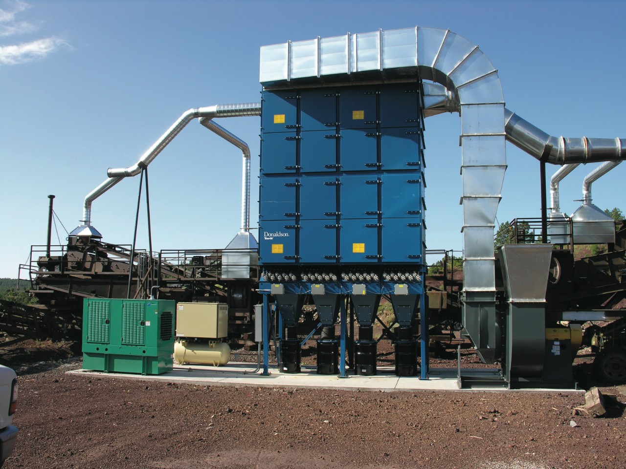 A centralized Donaldson Torit® Dalamatic® baghouse dust collector, with ducting to the dust-generating points at conveyor transfer points. Since this style of baghouse cleans its filters with compressed air, the facility needed to install an air compressor (green apparatus) and a compressed air dryer (tan equipment) beside the collector.